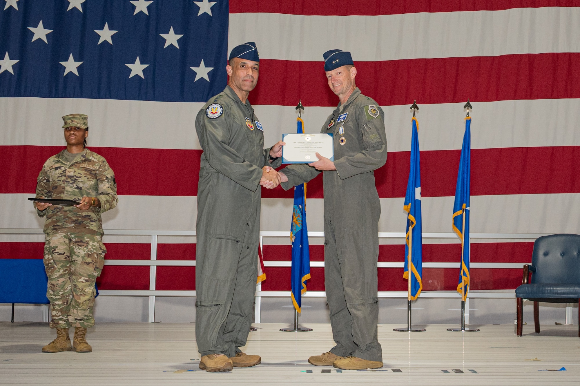 A general gives an award to another general on a stage with a large American flag in the background.