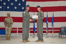 A general gives an award to another general on a stage with a large American flag in the background.