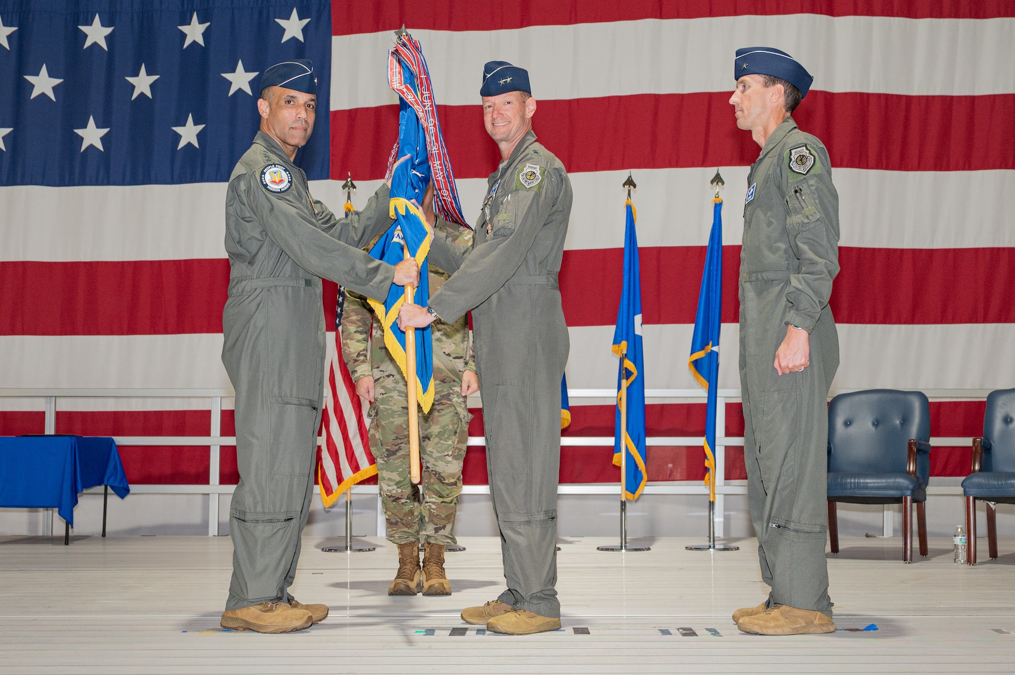 A general passes a guidon to another general on a stage with a large American flag in the background.