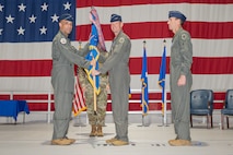 A general passes a guidon to another general on a stage with a large American flag in the background.