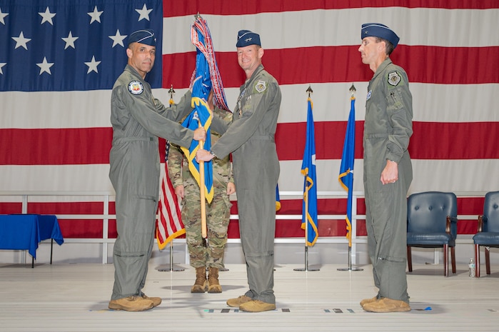 A general passes a guidon to another general on a stage with a large American flag in the background.