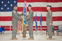 A general passes a guidon to another general on a stage with a large American flag in the background.