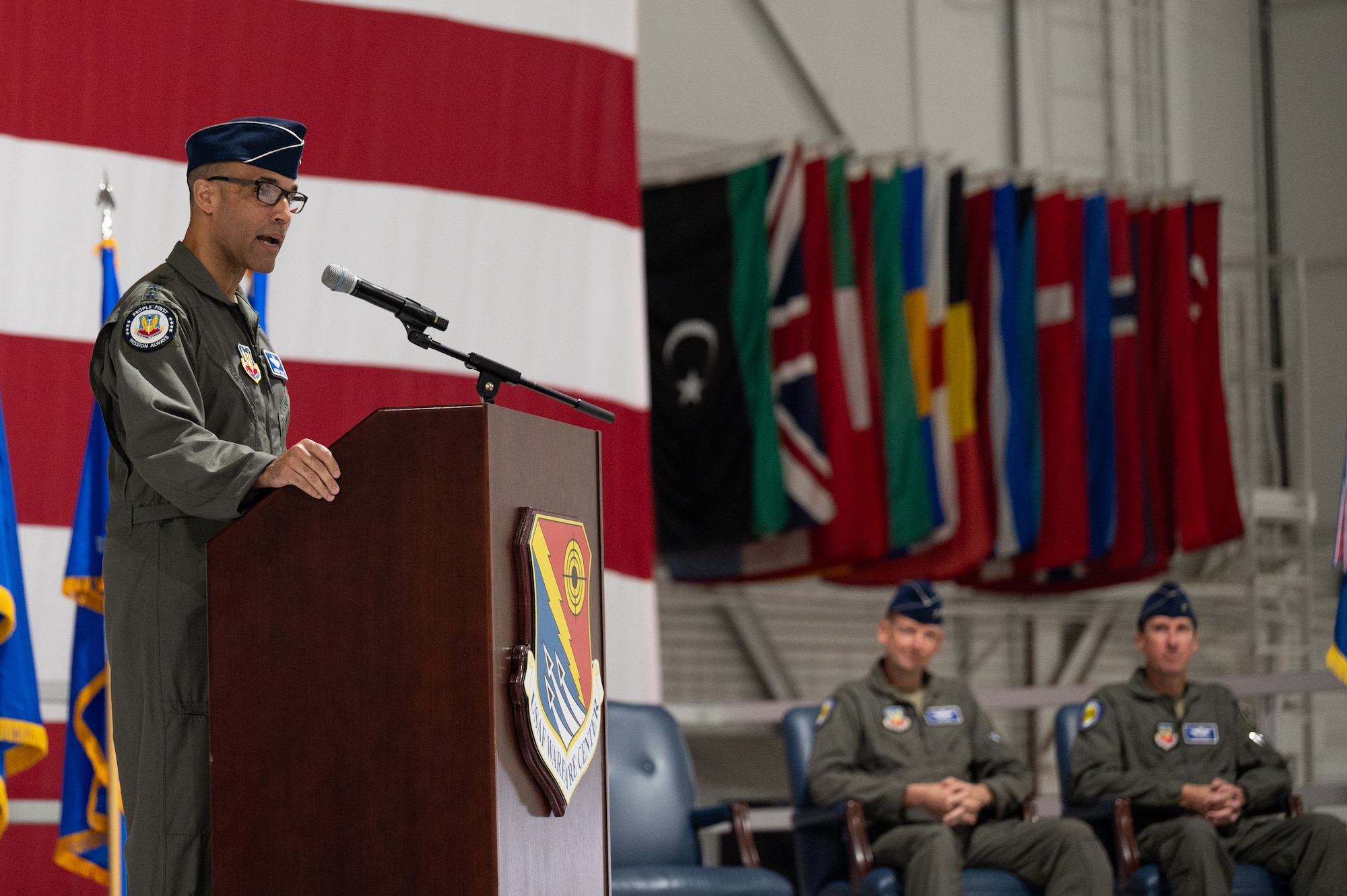 A military general speaks at a podium. There are two generals seated behind him, listening.