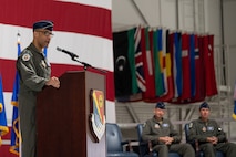 A military general speaks at a podium. There are two generals seated behind him, listening.