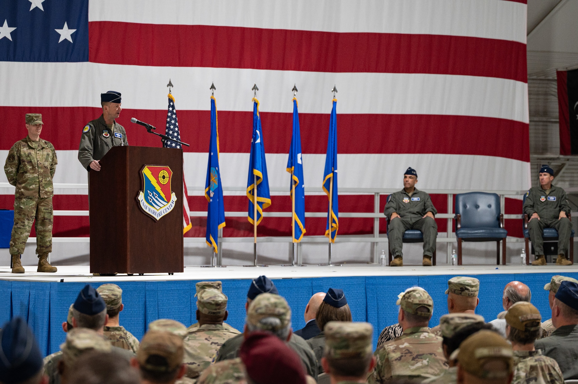 A military general speaks at a podium in front of a crowd with a large flag in the background.