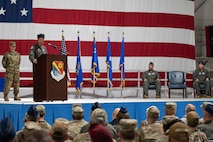 A military general speaks at a podium in front of a crowd with a large flag in the background.
