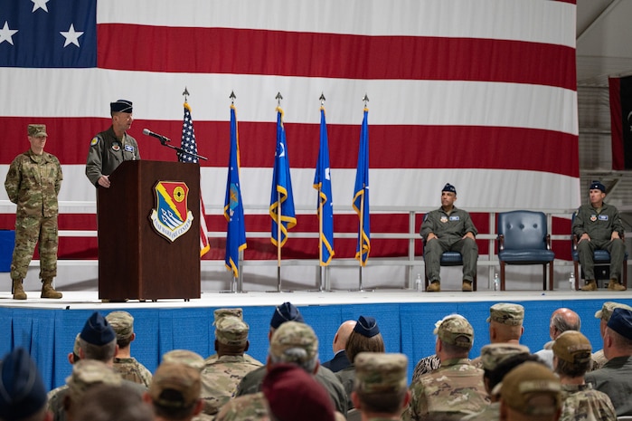 A military general speaks at a podium in front of a crowd with a large flag in the background.