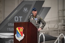 An Airman speaks at a podium. There is an F-35 in the background.