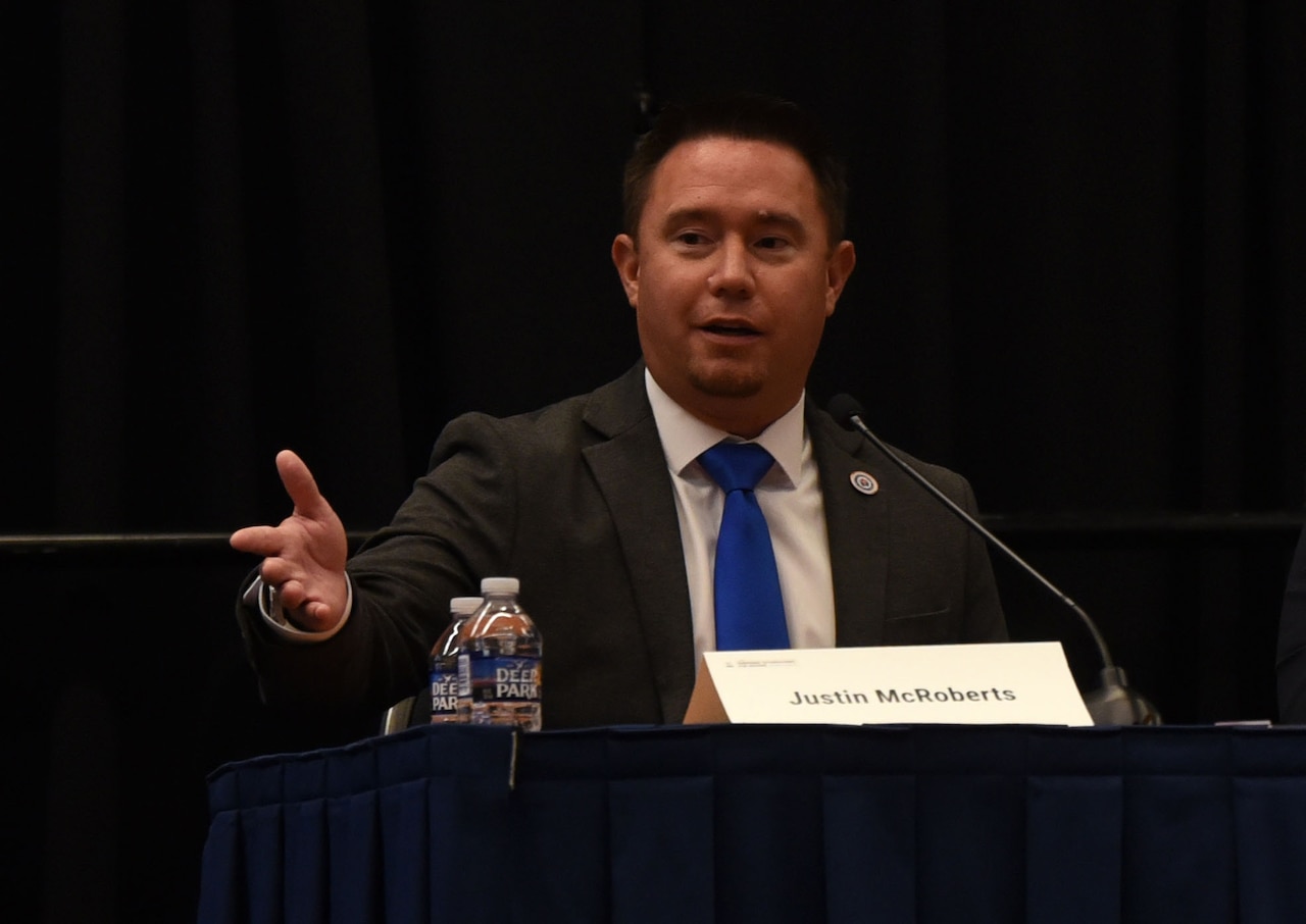 A person seated in a suit speaks to an audience.