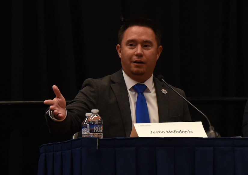A person seated in a suit speaks to an audience.