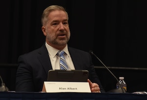 A person seated in a suit speaks to an audience.