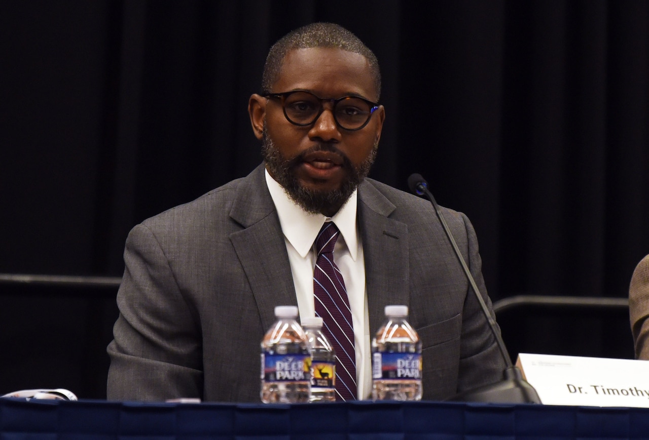 A person seated in a suit speaks to an audience.