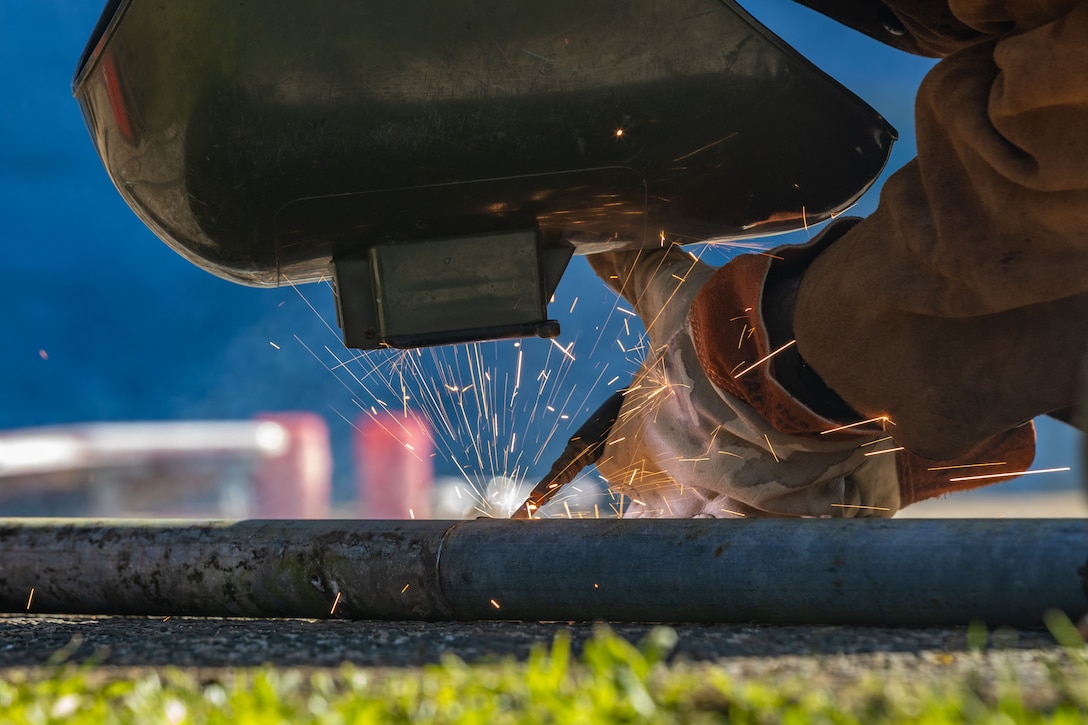 A sailor in a protective helmet and gloves welds something on the ground outdoors, sending sparks upward.
