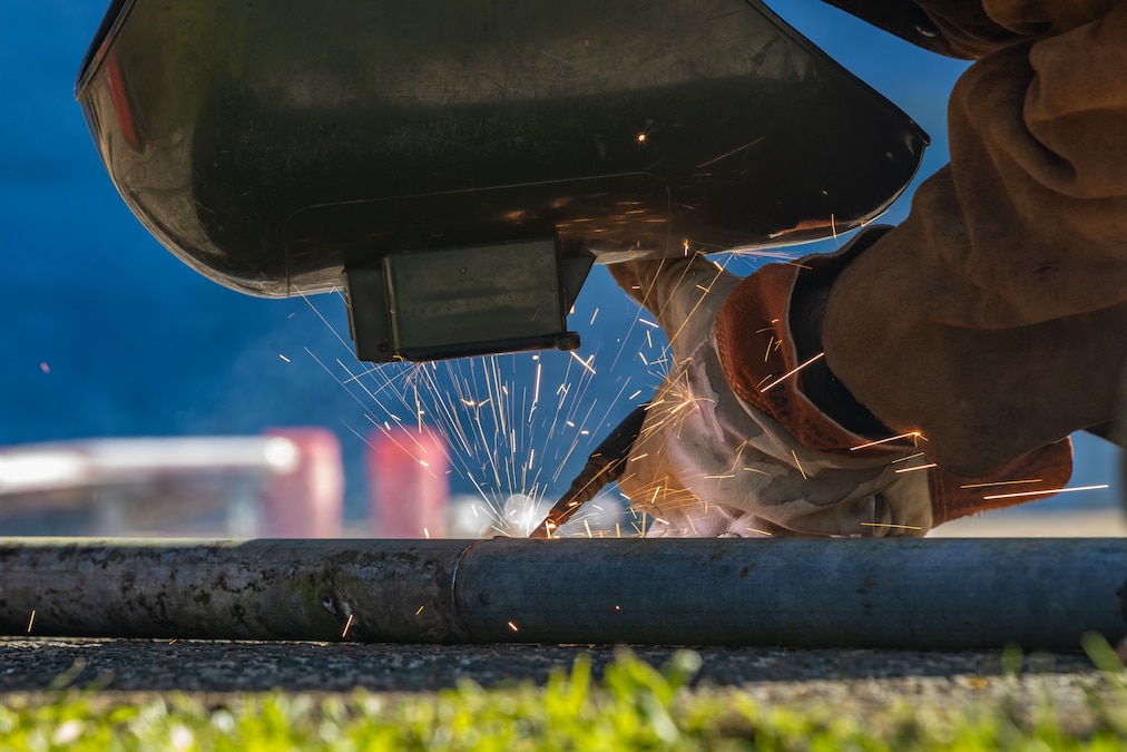 A sailor in a protective helmet and gloves welds something on the ground outdoors, sending sparks upward.