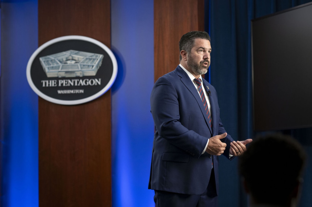 A man in a business suit is speaking in a room while looking to the right. Behind him is a sign on the wall that shows a five-sided building and reads “The Pentagon.”