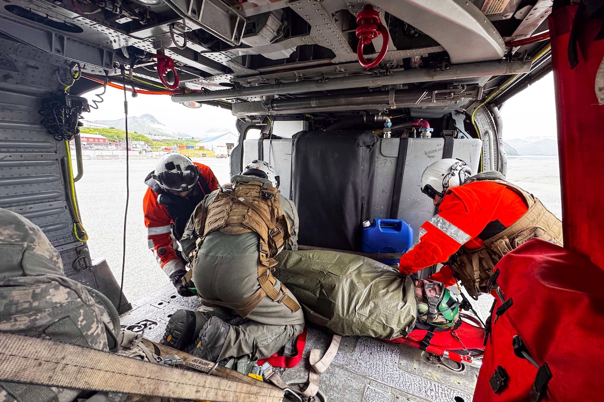 Two people in bright orange jackets, helmets and flight gear and one person in a flight suit with a helmet and a vest on secure a person on a stretcher to the inside of a helicopter.