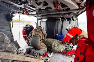 Two people in bright orange jackets, helmets and flight gear and one person in a flight suit with a helmet and a vest on secure a person on a stretcher to the inside of a helicopter.