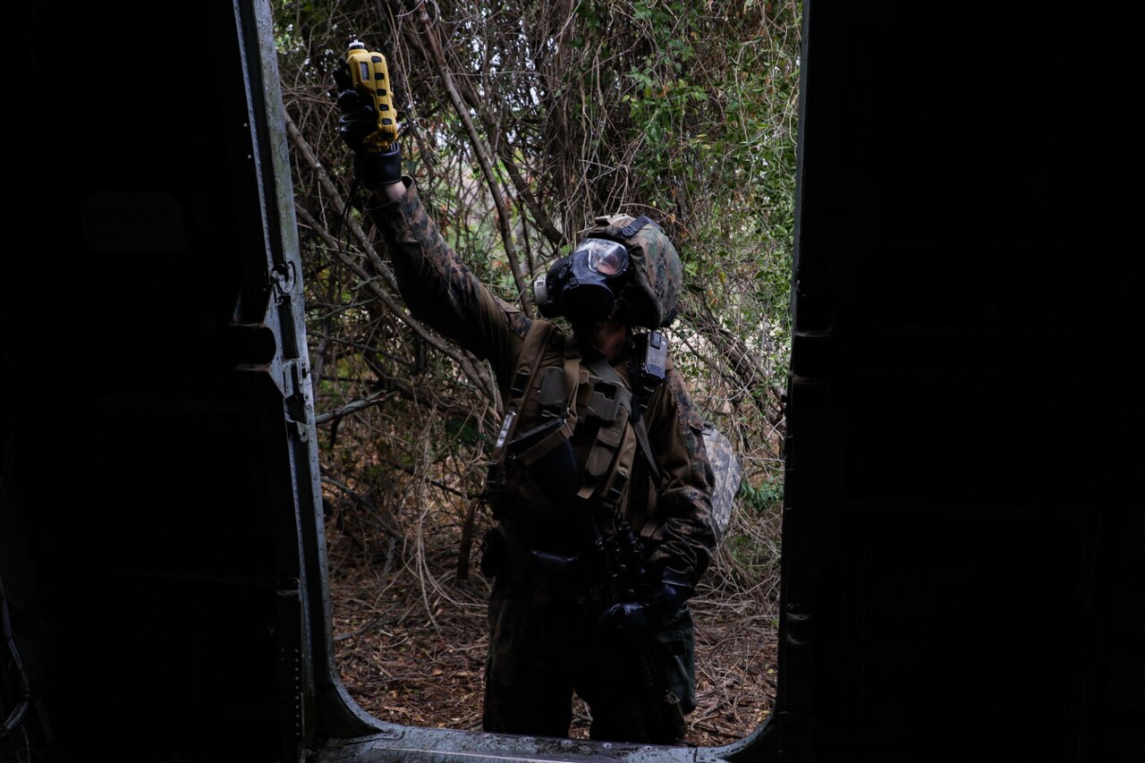 A person wearing camouflage personal protective equipment scans the door of an aircraft in a woodland environment.