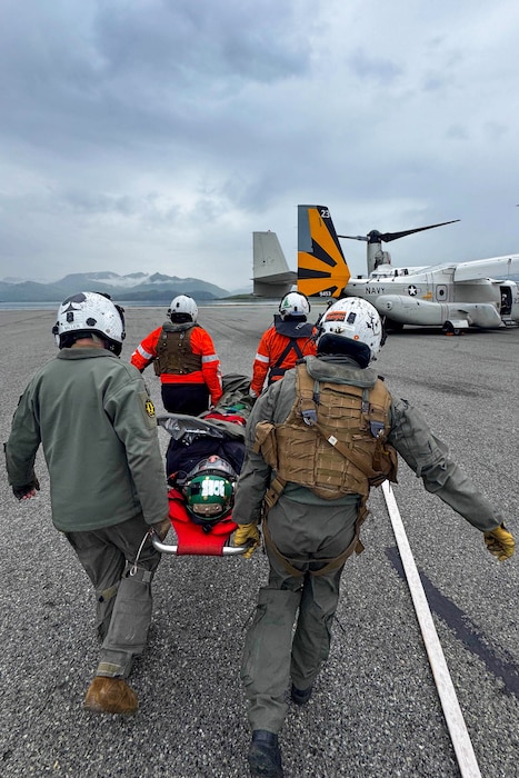 Two people in bright orange jackets and two people in flight suits and helmets carry a person on a stretcher toward a military aircraft. There are mountains in the background and clouds in the sky.