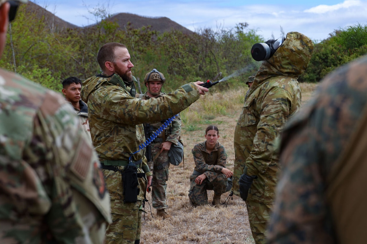 A person wearing a camouflage military uniform sprays a substance from a hose onto another person wearing camouflage personal protective equipment while a group of people stand and watch.