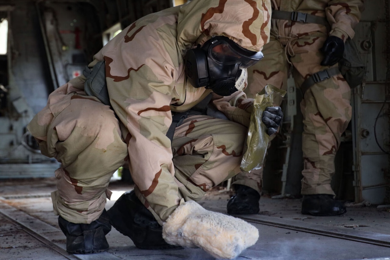A person wearing a camouflage personal protection equipment wipes a substance on the floor of an aircraft.