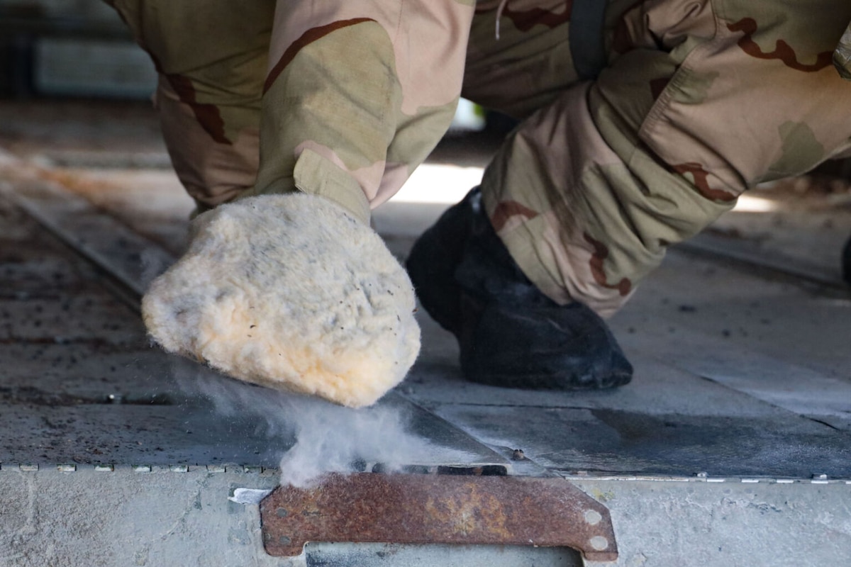 A close-up picture of the hand of a person wearing a camouflage personal protective equipment, wiping a substance on the floor of an aircraft. Smoke is rising from a rusted metal handle.