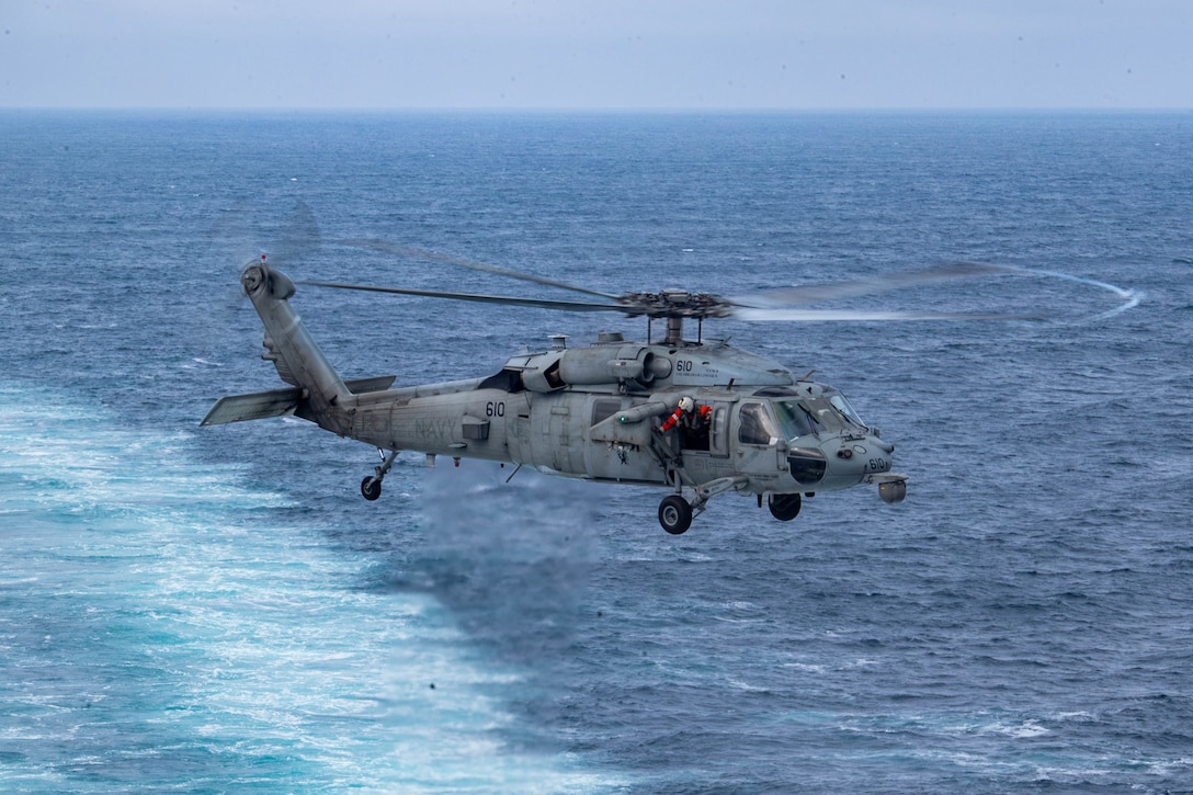 A helicopter hovers above the ocean while a sailor in red gear looks out of the open window on an overcast day.
