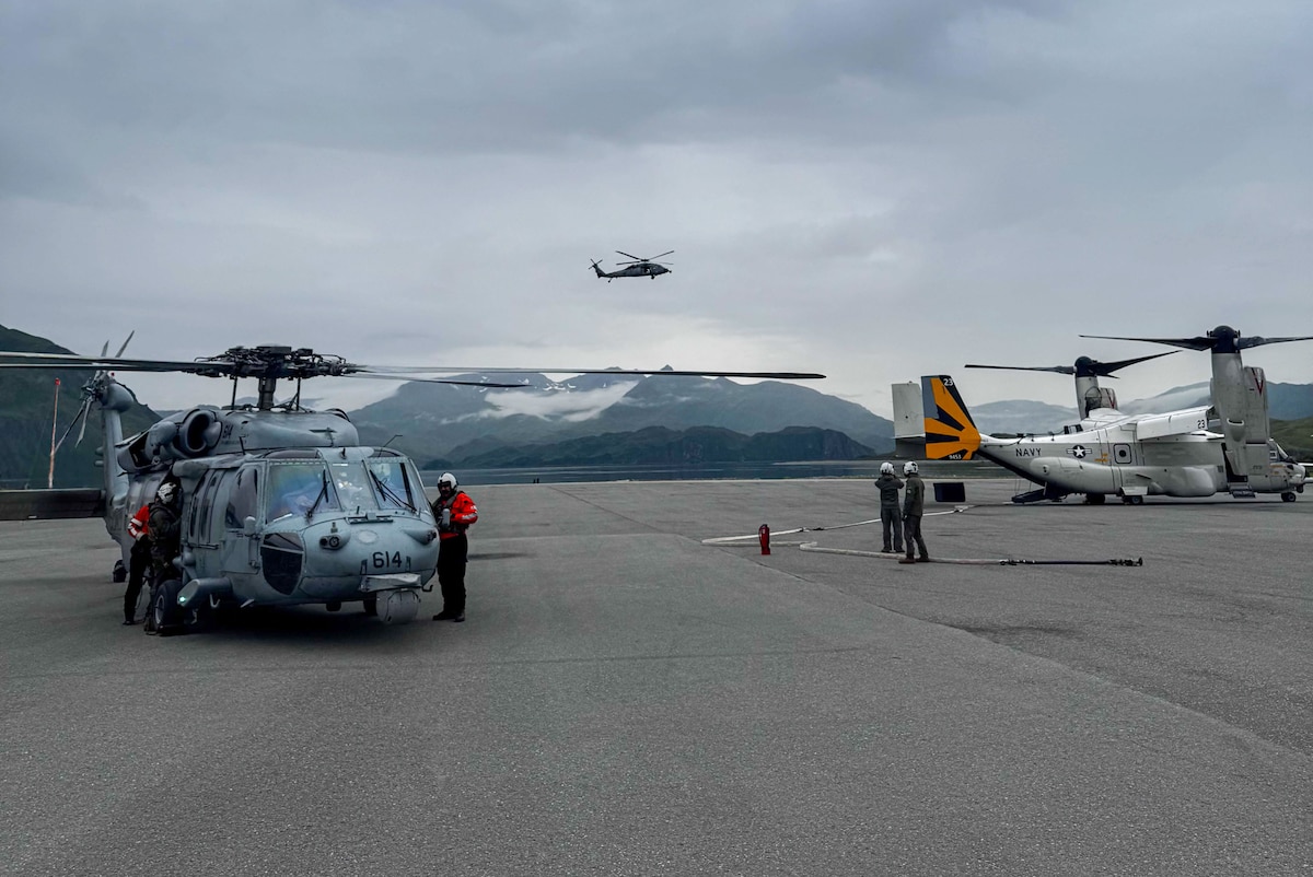 Two people in flight suits stand around a military helicopter while another two people in flight suits look at another military aircraft on the tarmac. There is another helicopter flying in the background.