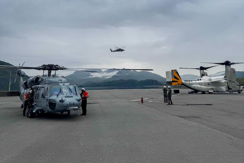 Two people in flight suits stand around a military helicopter while another two people in flight suits look at another military aircraft on the tarmac. There is another helicopter flying in the background.