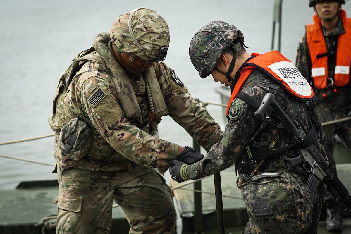 Two soldiers in camouflage military uniforms turn a bolt on a temporary bridge. The soldier on the right is carrying a gun on his back and wearing a bright orange life jacket. There is another soldier in similar attire in the background.