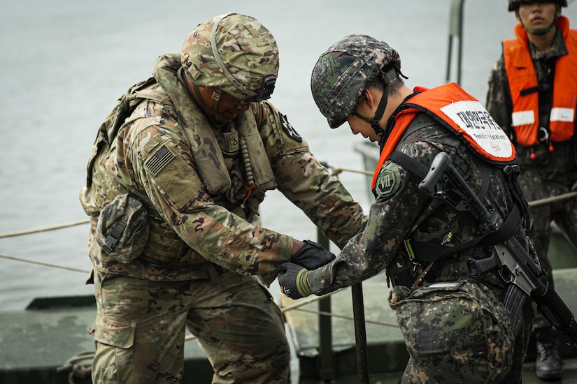 Two soldiers in camouflage military uniforms turn a bolt on a temporary bridge. The soldier on the right is carrying a gun on his back and wearing a bright orange life jacket. There is another soldier in similar attire in the background.