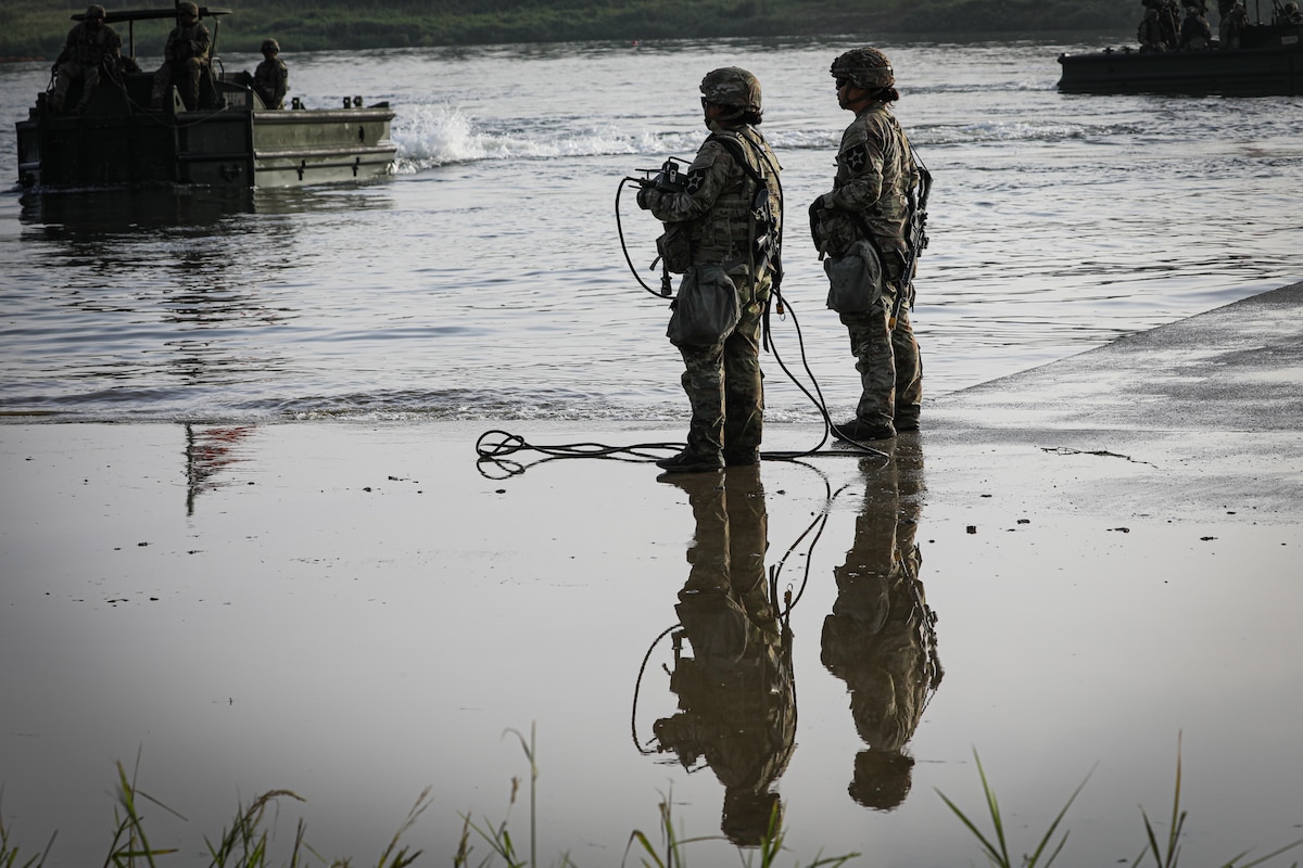 Two soldiers in camouflage military uniforms stand at the shallow side of a body of water while watching a small boat carrying other soldiers come toward them. The reflection of the soldiers standing in the water is shown in the foreground.