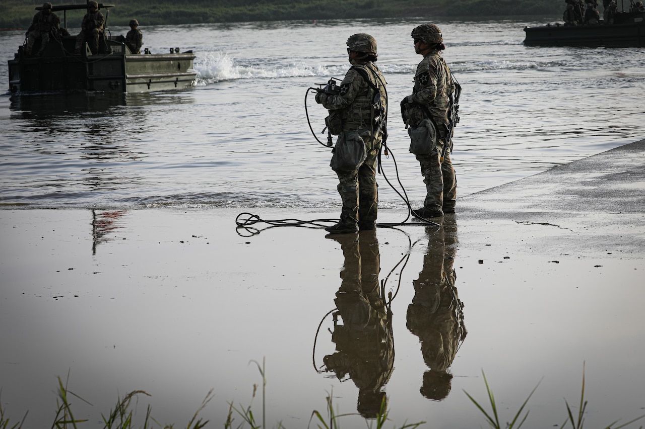 Two soldiers in camouflage military uniforms stand at the shallow side of a body of water while watching a small boat carrying other soldiers come toward them. The reflection of the soldiers standing in the water is shown in the foreground.