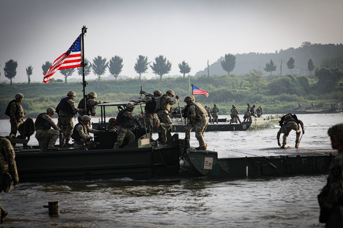 A group of soldiers in camouflage uniforms and helmets set up a temporary bridge across a river. There are more soldiers in similar attire on a boat in the background and two American flags flying from poles on each boat.