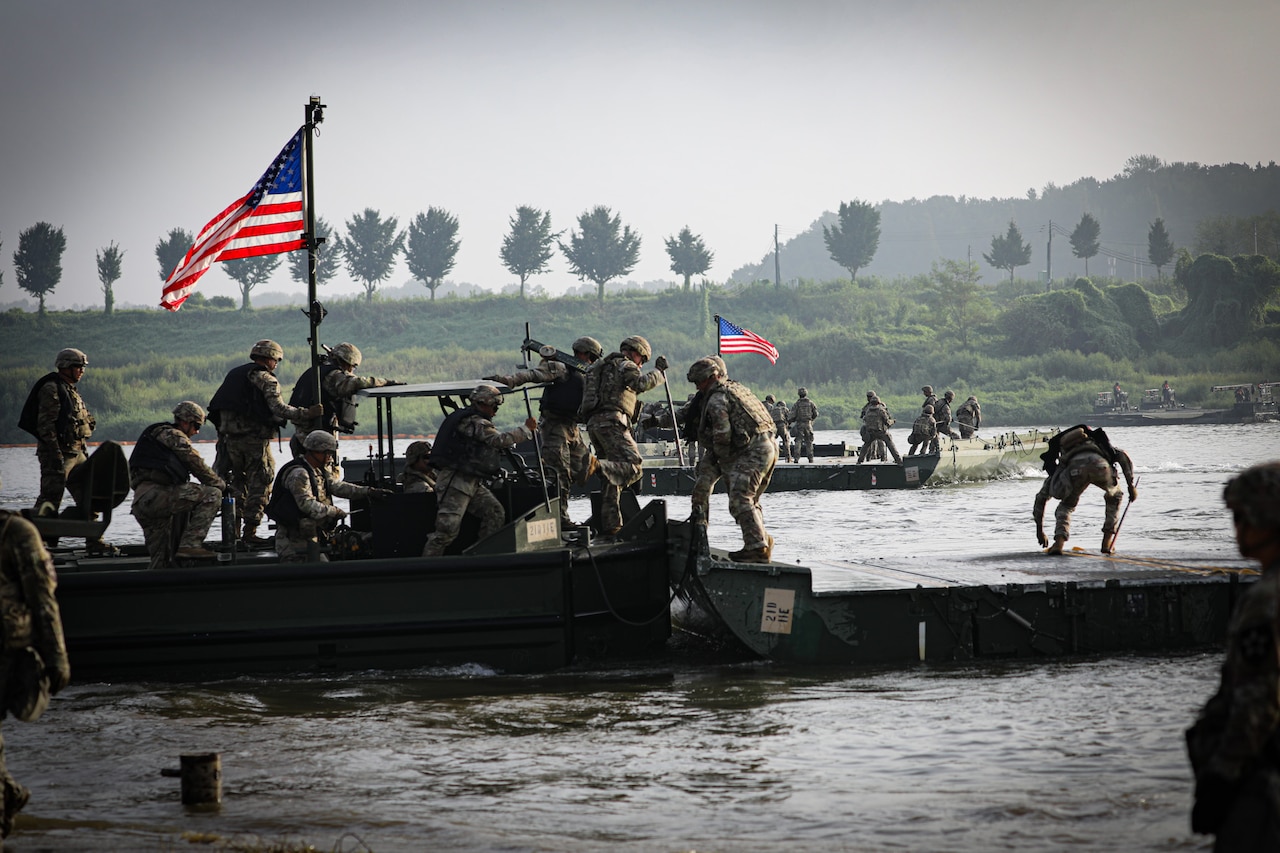 A group of soldiers in camouflage uniforms and helmets set up a temporary bridge across a river. There are more soldiers in similar attire on a boat in the background and two American flags flying from poles on each boat.