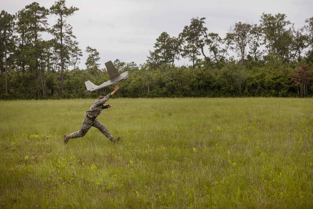 A Marine runs in a field while launching an unmanned aerial system into a gray, cloudy sky.