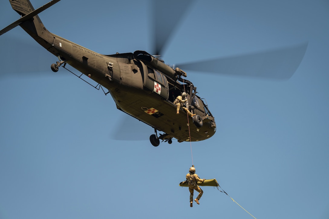 A soldier holding a stretcher is hoisted to an airborne helicopter against a blue sky.