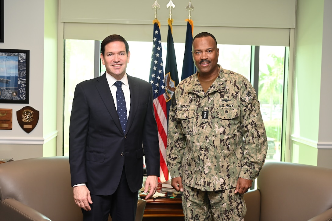 A man in a black suit and a man in a camouflage uniform pose for a photo in front of flags.