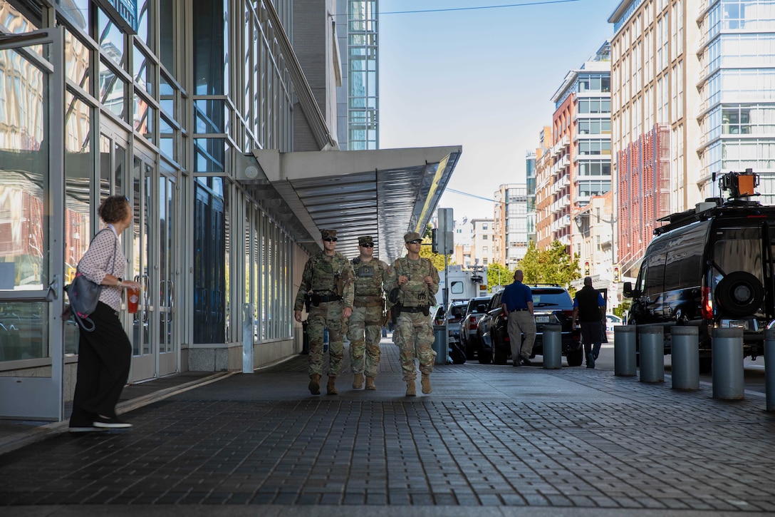 Three guardsmen walk down a city street next to a building while a person to the left watches and cars are parked to the right.