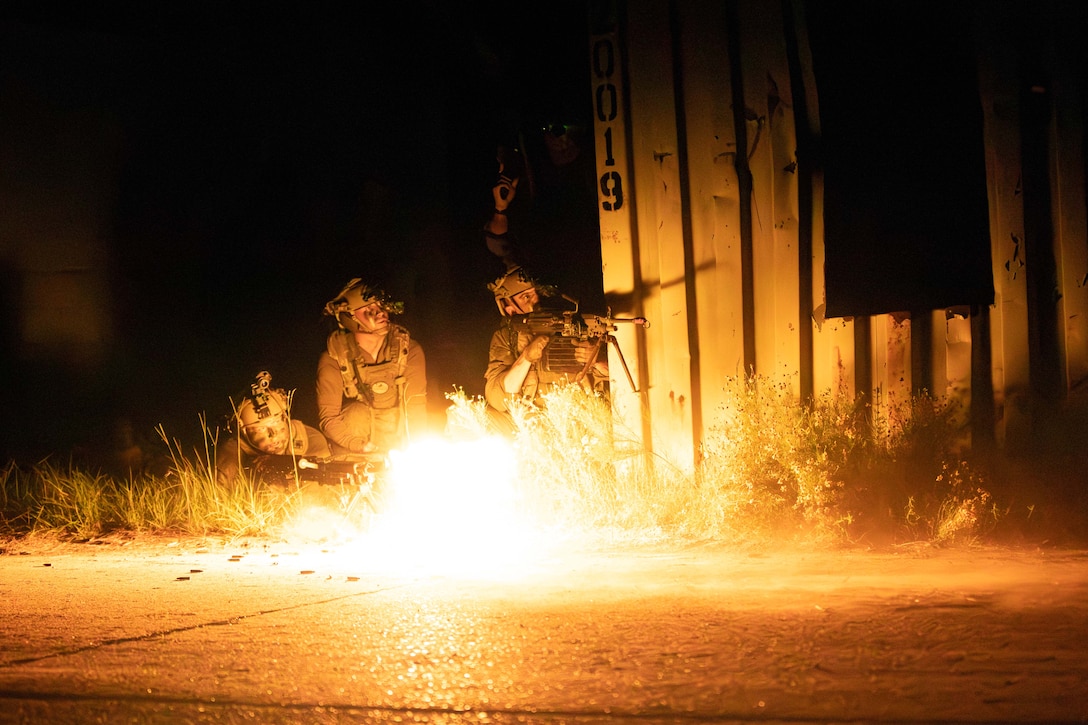 Three soldiers in tactical gear crouch near a metal wall in the grass next as one fires a service weapon, illuminating the scene with yellow fire.