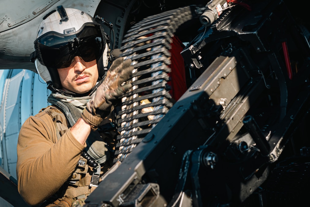A sailor in a helmet loads a magazine into a machine gun while sitting in a parker aircraft during the day.