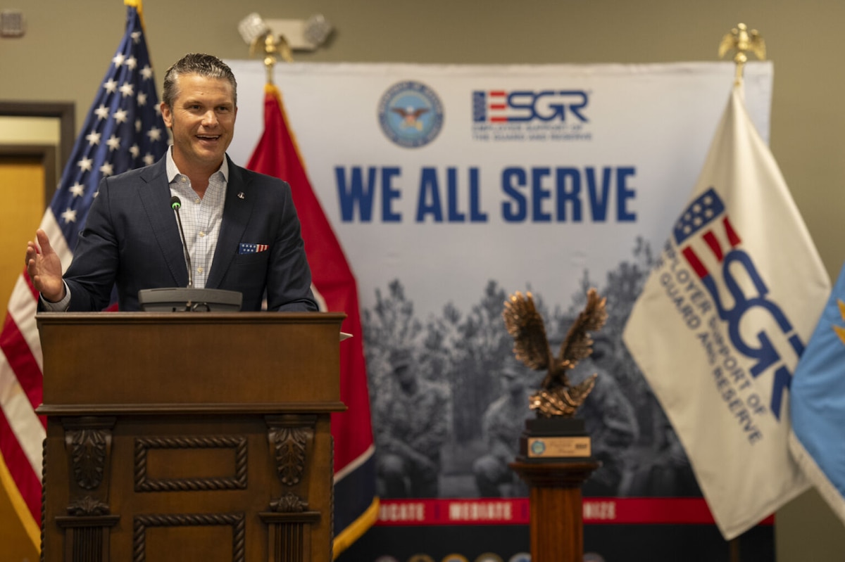 A man in business attire stands speaking behind a podium. In front of him is a large trophy fashioned in the shape of a flying eagle and behind him is a U.S. flag and a sign that reads “We All Serve.”