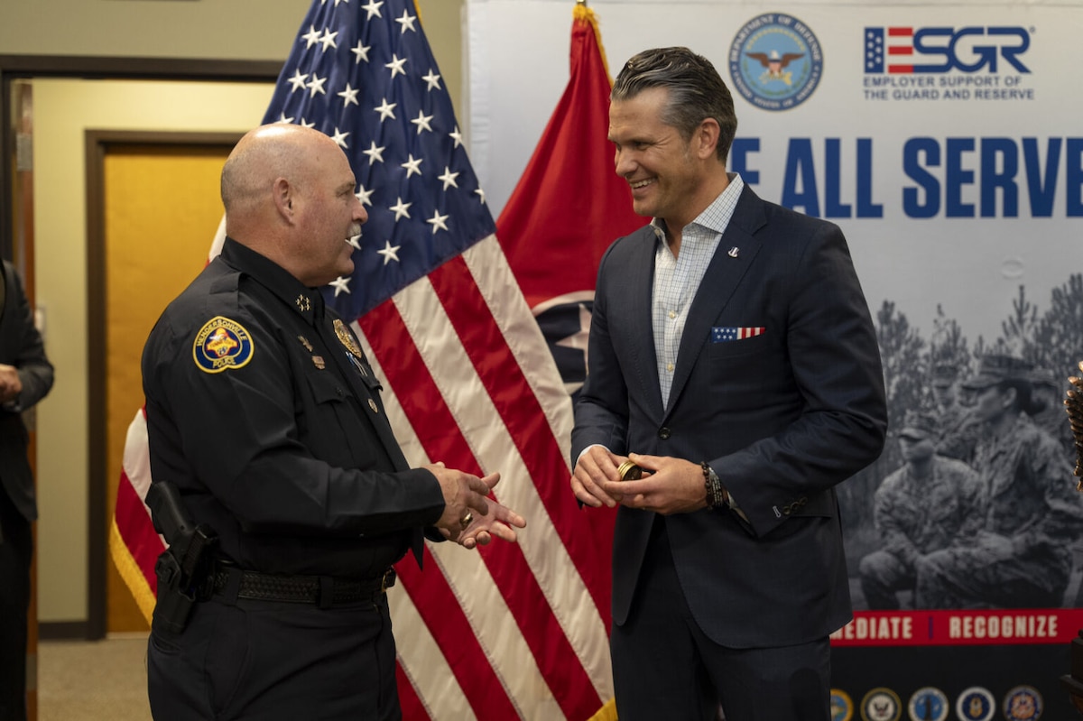 Two men, one in a police officer’s uniform and the other in civilian business attire, are talking. There is a U.S. flag behind them.