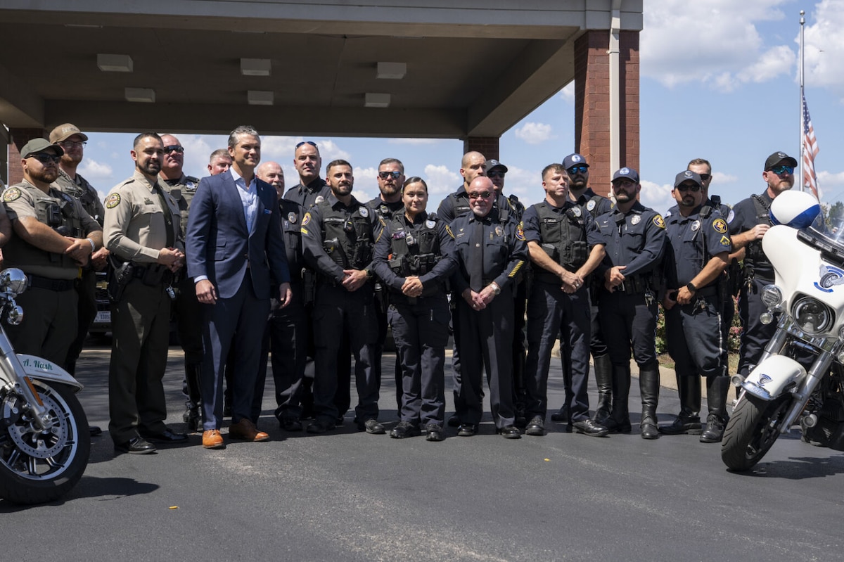 A man in civilian business attire stands amid roughly 20 men in police officer uniforms. They are flanked on either side by a pair of police motorcycles.