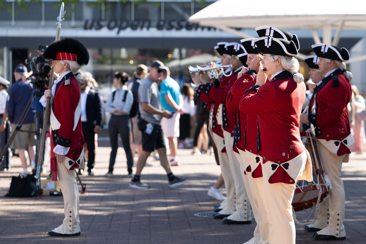 People dressed in colonial period uniforms stand in formation and play instruments at an outdoor event.