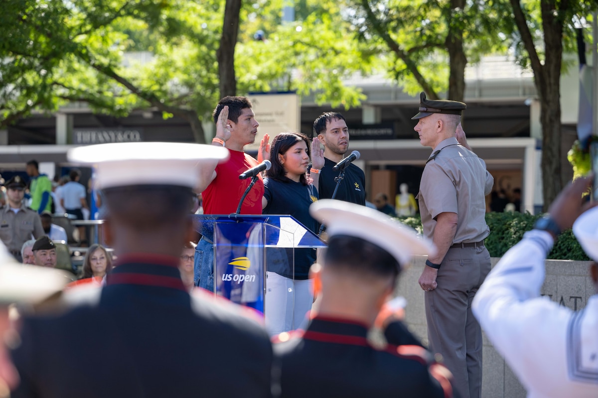 Three people stand and speak with their right hand raised as they face a man wearing a formal military uniform who also stands with his right hand raised.