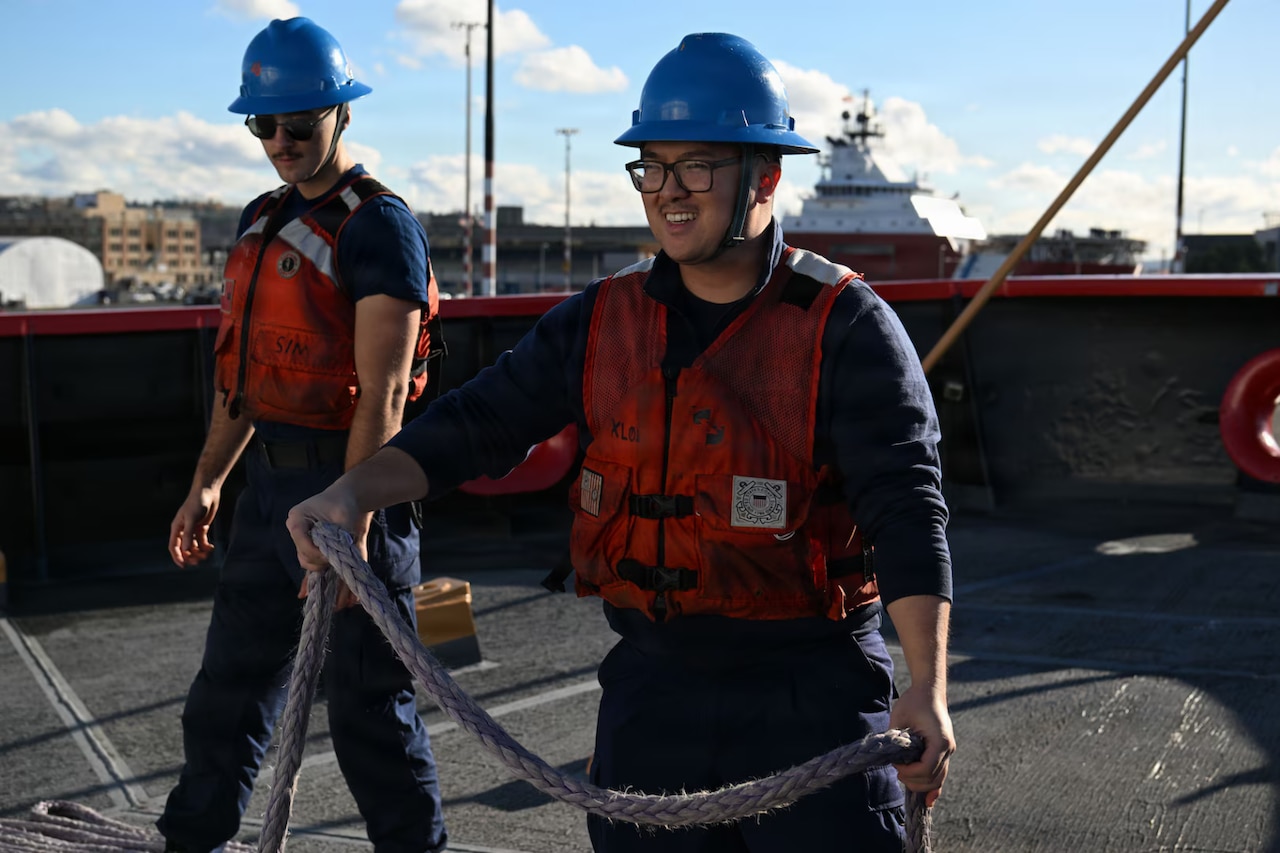 Two service members wearing life jackets and hard hats pull a large rope on a ship.