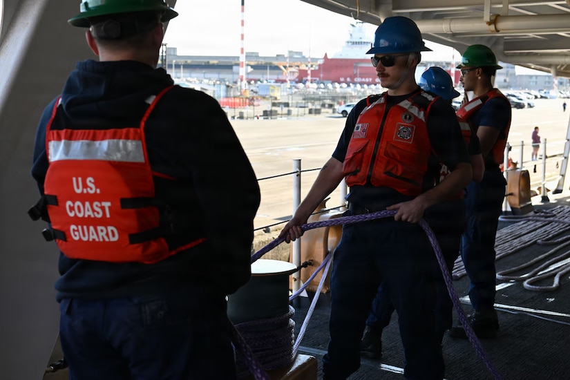 Two service members wearing life jackets and hard hats pull a large rope on a ship.