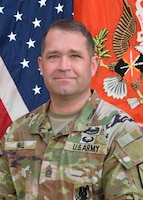 Photo of a soldier in camouflaged utility uniform with an American flag on the left and a Signal Regiment flag on the right.