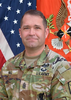 Photo of a soldier in camouflaged utility uniform with an American flag on the left and a Signal Regiment flag on the right.